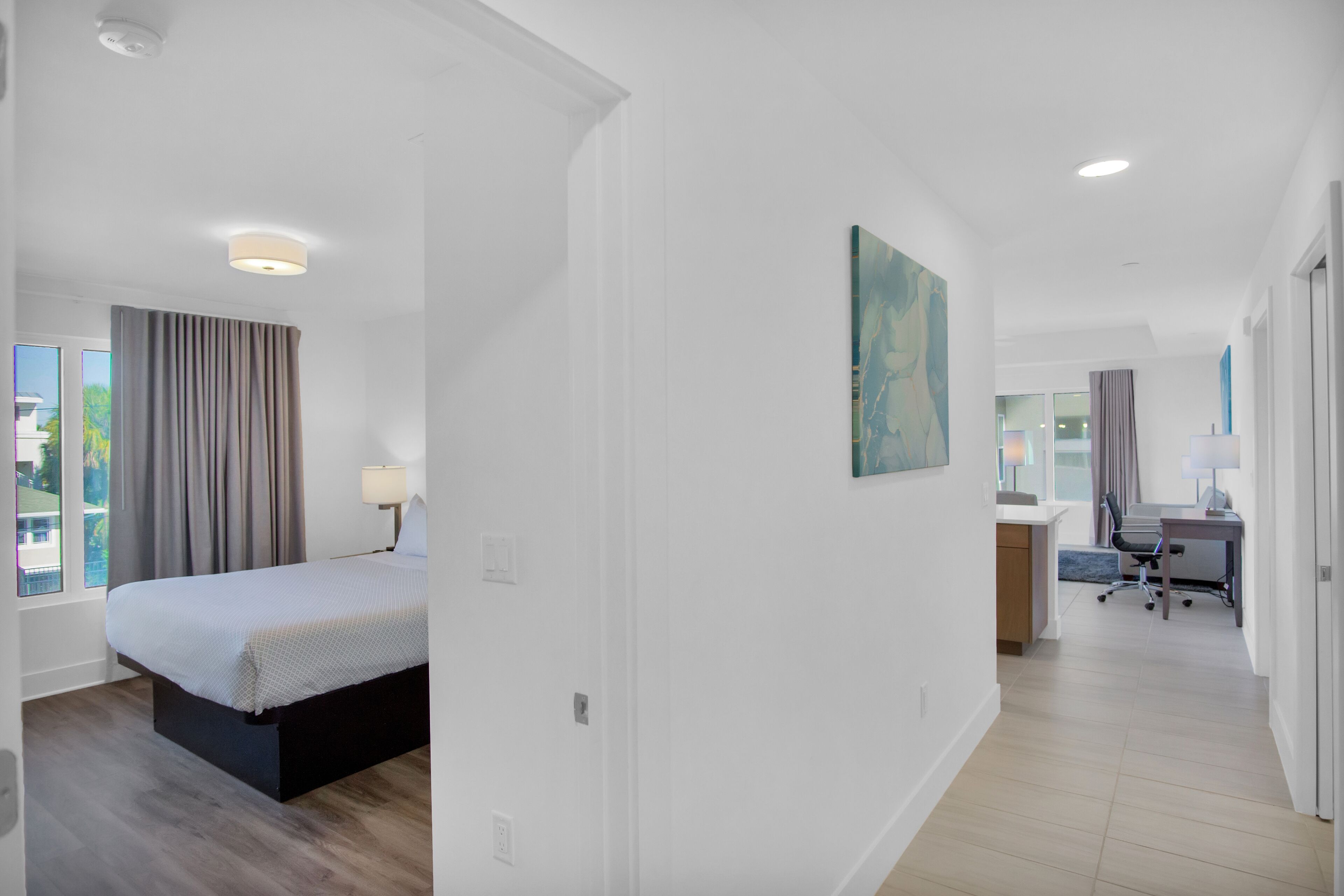 Une chambre d’hôtel moderne, équipée d’un lit, d’un bureau et d’une chaise, offrant une vue sur l’extérieur et ornée d’un tableau au mur.