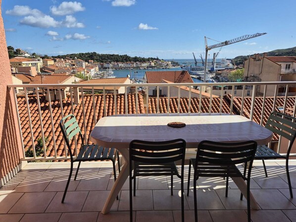 A balcony with a table and chairs, overlooking a coastal town with terracotta rooftops and a marina.