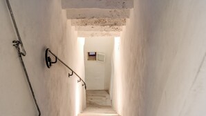 Un escalier étroit, muni d’une main courante sur le côté gauche, mène à une pièce dotée d’une petite fenêtre et d’une porte en haut.
