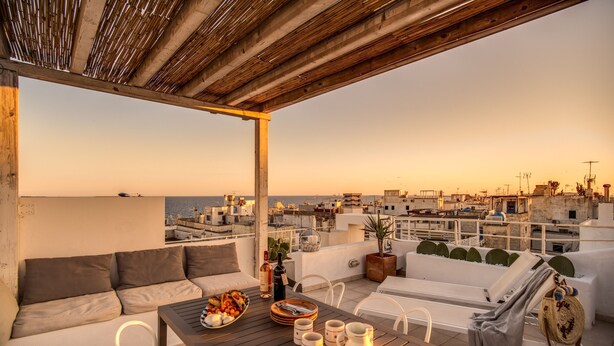 Une terrasse sur le toit agrémentée d’une pergola en bois, de mobilier d’extérieur blanc, d’une table à manger avec des assiettes et des tasses, et offrant une vue sur le paysage urbain.