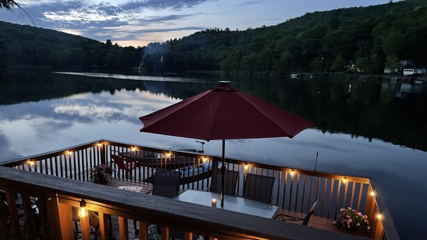 Une terrasse en bois avec une table et des chaises, entourée de guirlandes lumineuses, donnant sur un lac paisible, avec une colline boisée en arrière-plan.