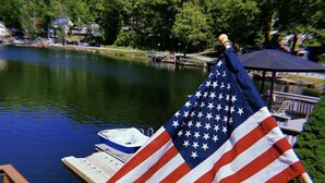 Un drapeau américain flotte sur un quai au bord d’un lac, avec un bateau et un kiosque en arrière-plan.