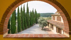 A courtyard with a brick archway, a pathway lined with cypress trees, and a building with a tiled roof. - Trilo standard first floor (Riparbella)