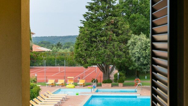 A swimming pool with lounge chairs and a tennis court in the background. - Trilo standard first floor (Riparbella)