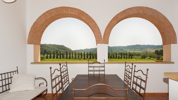 A room with a bed, a table, and two arched windows offering a view of a vineyard and hills. - Bilocali Superior first floor terrace (Riparbella)