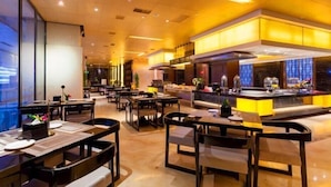 A modern restaurant interior with multiple dining tables and chairs, a central kitchen area, and large windows providing natural light. - Celebrity Hotel (Baokang)