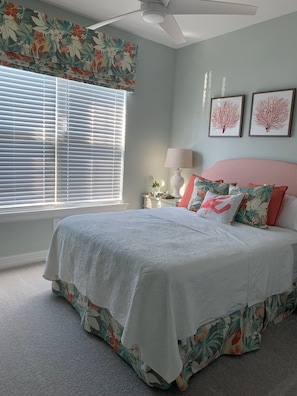 Une chambre à coucher avec un lit, des rideaux à motifs floraux, un ventilateur de plafond et deux tableaux encadrés accrochés au mur.