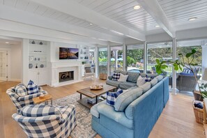Un salón espacioso con chimenea, muebles a cuadros azules y blancos, y un ventanal con vistas a un jardín.