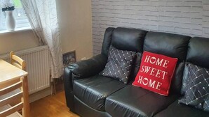 A living room with a black leather sofa, a red cushion with "HOME SWEET HOME" text, a brick-patterned wall, and a window with curtains.