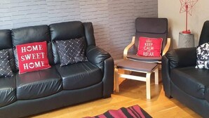 A living room with two black leather sofas, one with a red pillow that says "HOME SWEET HOME" and another with a red pillow that says "KEEP CALM AND RELAX". There is a wooden coffee table in the center, a red star-shaped floor lamp, and a framed picture on the wall.