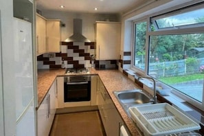 A modern kitchen with a central island, a built-in oven, a stainless steel sink, and a window with a view of greenery. - Lovely 4-bed Apartment in Harrow (Harrow)