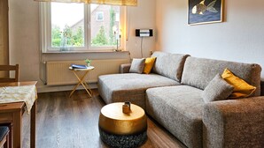 Un salón acogedor con un sofá gris, una mesa de centro de madera y una mesita redonda. Una ventana con cortina amarilla deja entrar luz natural y un cuadro enmarcado cuelga en la pared.