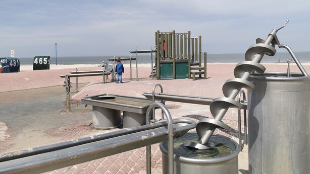Un baño público en la playa con una escalera de caracol metálica, un contenedor de basura y una persona cerca de la entrada.