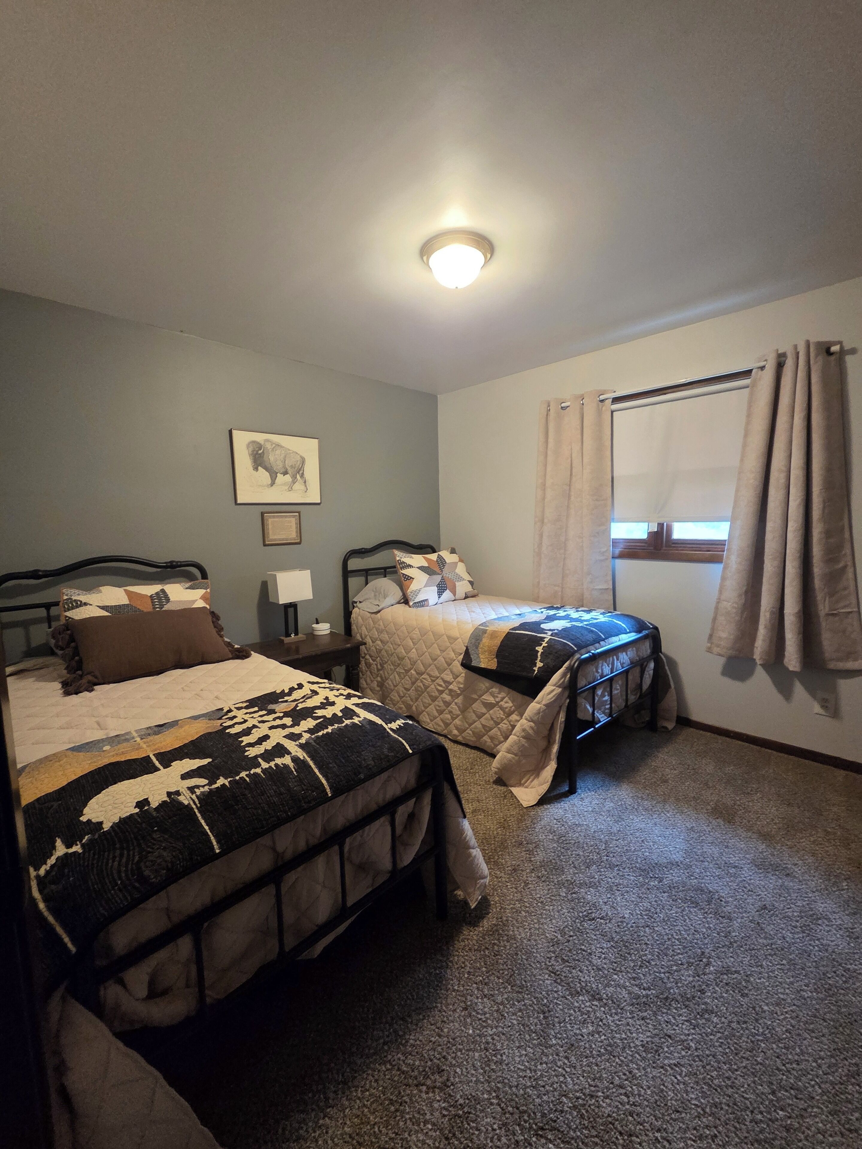 A bedroom with two beds, a window with curtains, a ceiling light, and a wall-mounted picture.