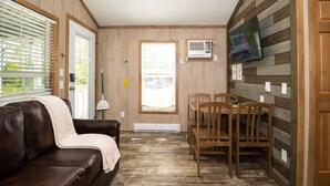 A cozy living room with a leather couch, wooden dining table, and chairs. There are two windows with blinds, a wall-mounted TV, and a ceiling fan.