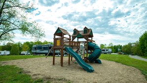 A playground with a slide and climbing structures, surrounded by sand and greenery, with RVs parked nearby.
