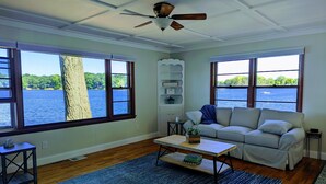 A spacious living room with large windows overlooking a lake, a ceiling fan, and a striped sofa.