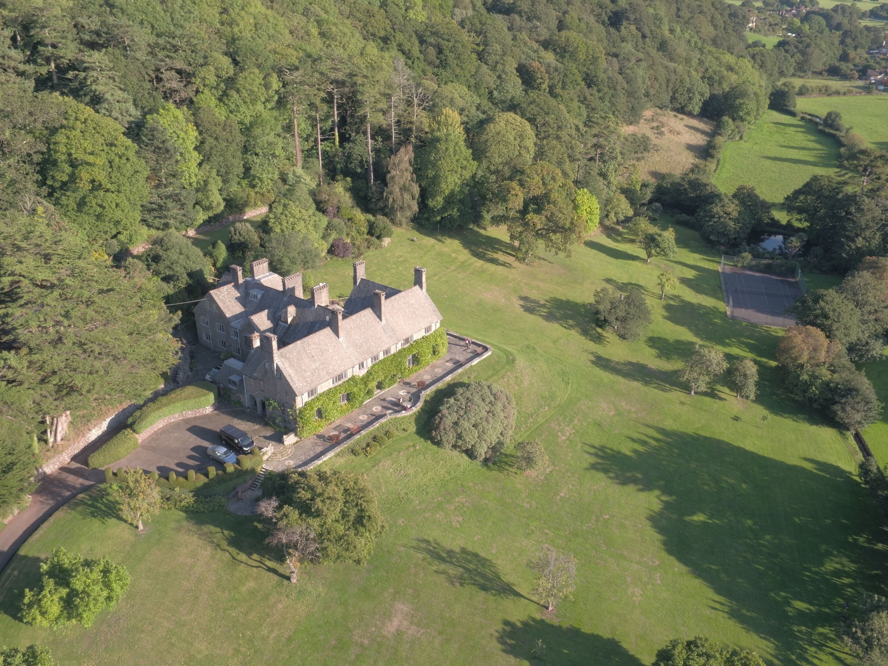 Une vue aérienne d’un grand bâtiment historique en pierre, entouré de vastes pelouses verdoyantes et d’une forêt dense.