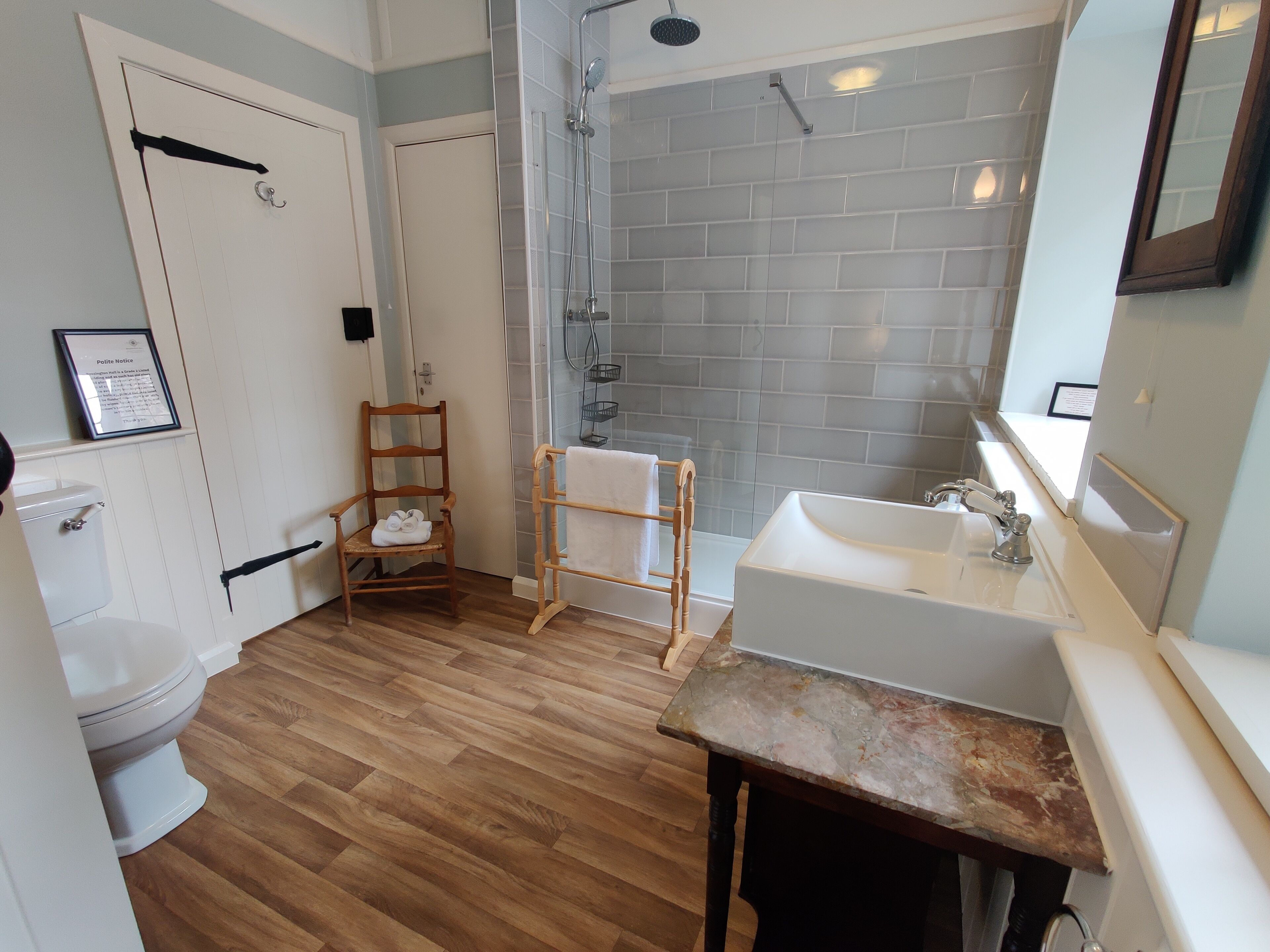 Un baño con suelo de madera, un lavamanos blanco, una ducha y una silla de madera.