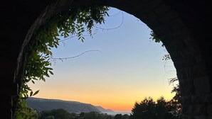 Une fenêtre en arc surmontée de pierres, offrant une vue sur le coucher du soleil, avec de la verdure et des bâtiments visibles au loin.