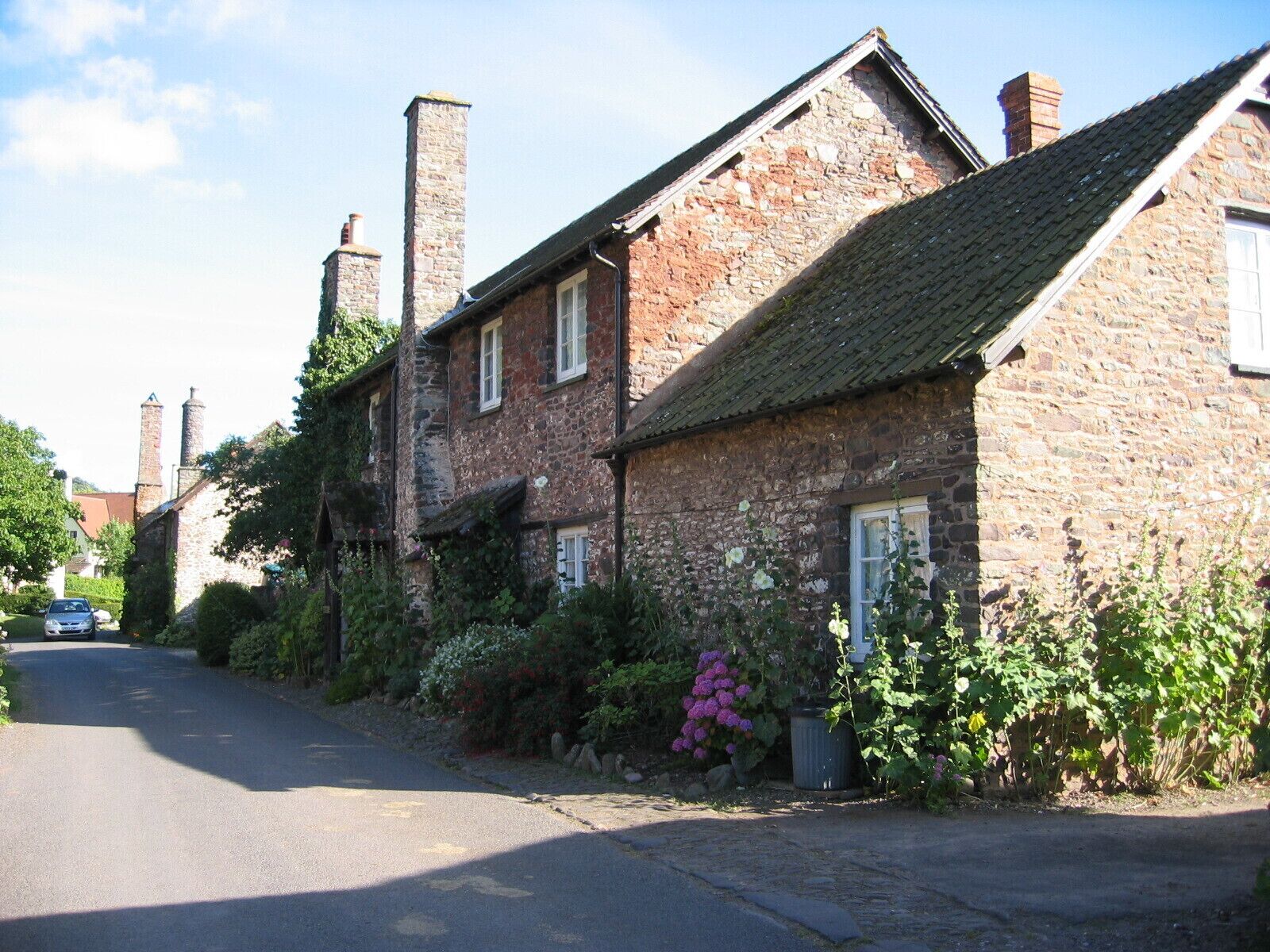 A traditional stone building with a sloped roof and chimneys, surrounded by lush greenery and flowering plants.