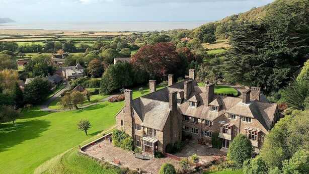 Un vaste manoir en pierre, doté de nombreuses cheminées, entouré de champs verdoyants et d’arbres luxuriants.