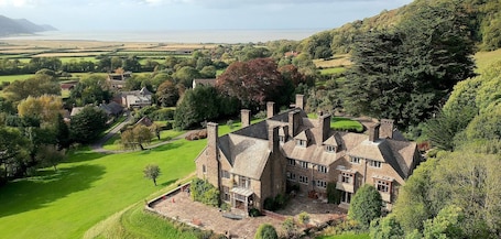 A large stone manor with multiple chimneys, surrounded by lush green fields and trees.. Bossington Hall