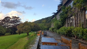 Un chemin de pierre mène à des bancs et des tables en bois, le tout étant entouré d’une végétation luxuriante et d’un bâtiment en pierre doté de grandes fenêtres.
