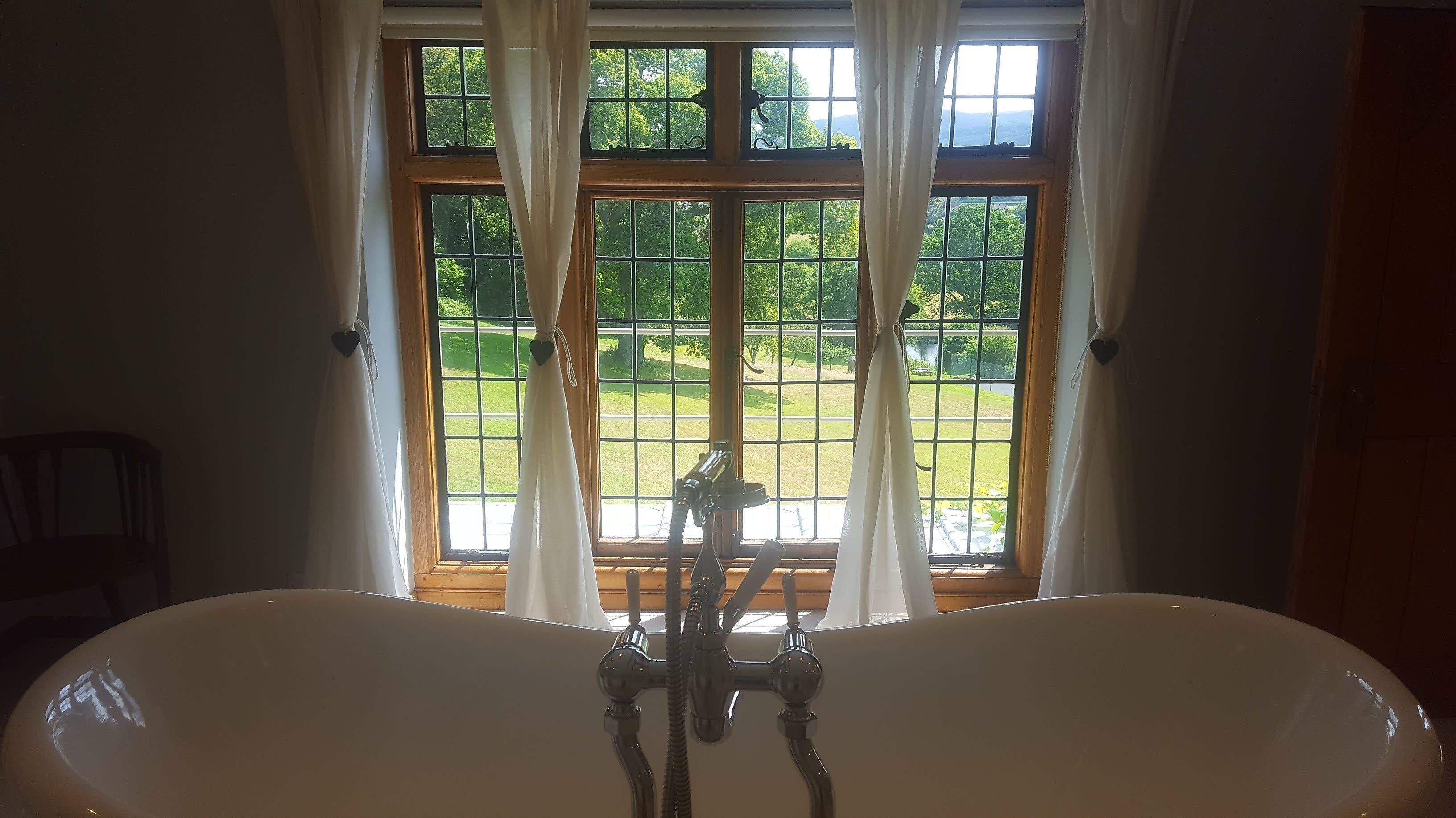Een badkamer met een vrijstaand bad, een raam met gordijnen en uitzicht op een groen landschap.