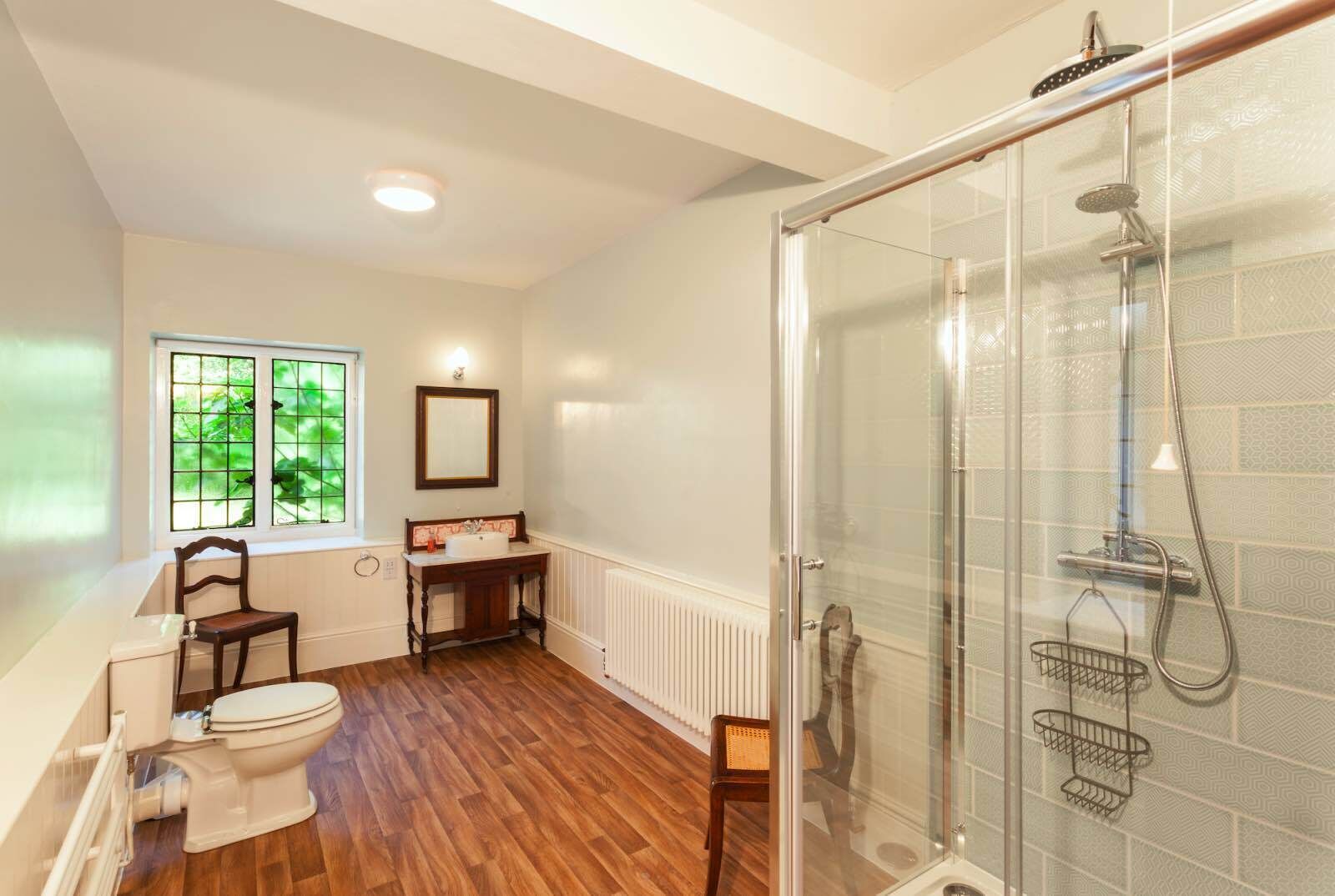 A bathroom with a glass-enclosed shower, a toilet, a vanity with a mirror, and a window with greenery outside.