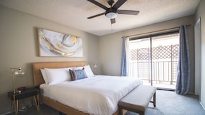 Un dormitorio con cama, mesitas de noche, ventilador de techo, un ventanal con cortinas y vistas a un balcón.