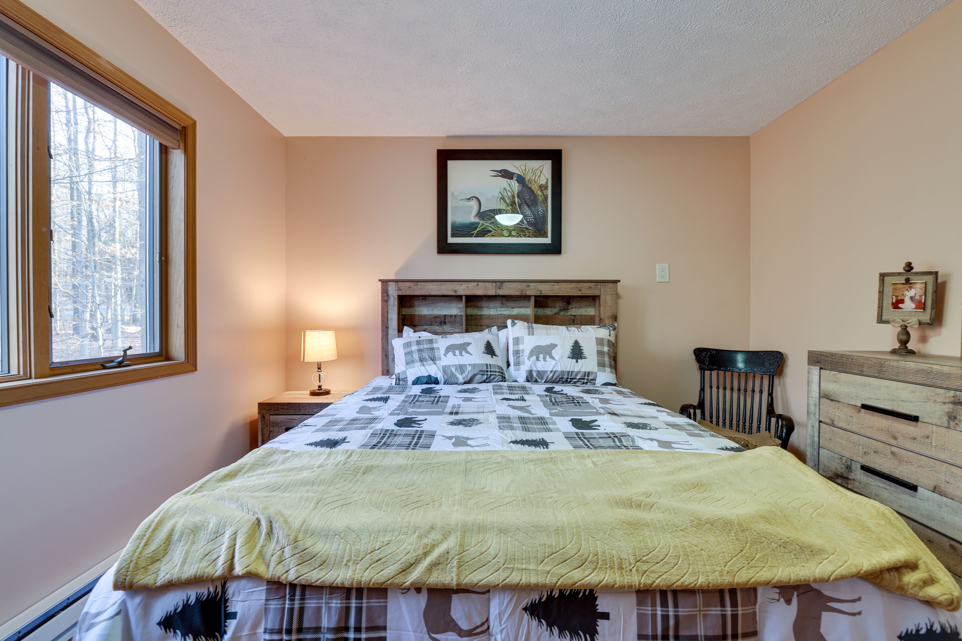 Un dormitorio con cama, mesita de noche, cómoda, ventana y un cuadro en la pared.
