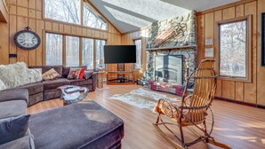 A spacious living room with a stone fireplace, wooden walls, a large TV, and a cozy seating area. - Large rustic style home in gated community: Fire Pit - Lake - Beach - pools! (Gouldsboro)