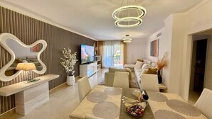 A modern living room with a dining area, featuring a large TV, a sofa set, and a unique wall decor. - Casares Beach Doña Julia Golf Apartment With Private Garden Direct Pool Access (Casares)