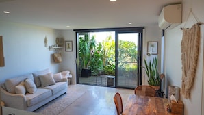 Ein modernes Wohnzimmer mit einer Couch, einem Esstisch und Stühlen. Eine Glasschiebetür führt zu einem Balkon mit Pflanzen.