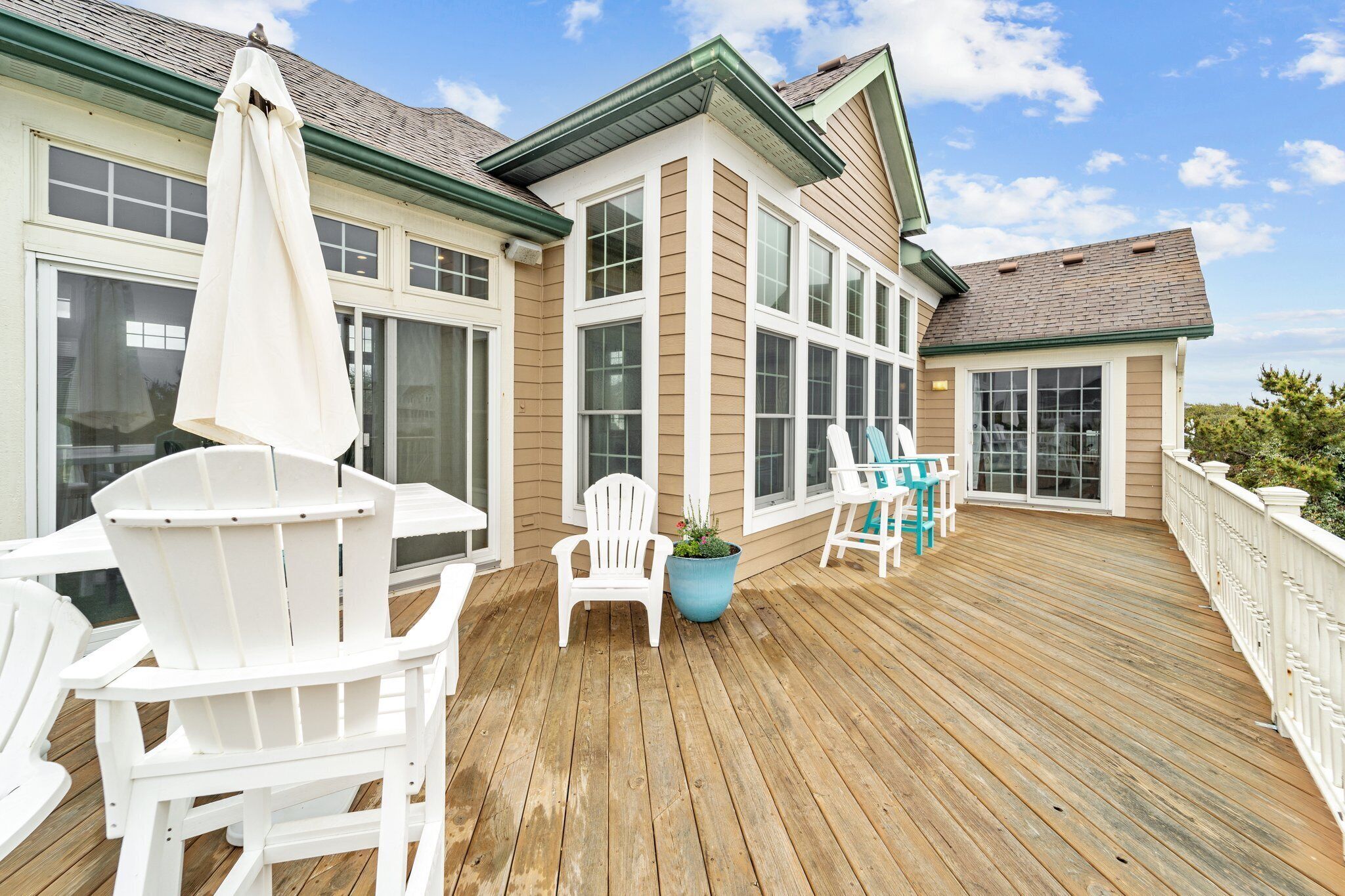 A wooden deck with white chairs and a large umbrella, a potted plant, and a view of a house with large windows and a green roof.