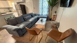 Un salon avec un canapé, deux fauteuils, une table basse, un téléviseur et, au fond, une cuisine.
