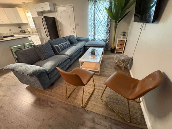 A living room with a sofa, two chairs, a coffee table, a TV, and a kitchen in the background.