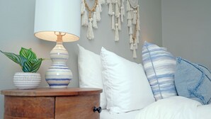 Une chambre à coucher bien rangée, avec une table de chevet en bois, une lampe, une plante en pot et un lit agrémenté d’oreillers à rayures blanches et bleues.