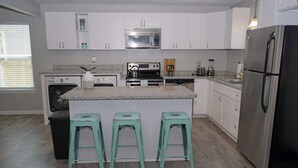 Une cuisine moderne avec des armoires blanches, un réfrigérateur en acier inoxydable, un four à micro-ondes et une cuisinière. On trouve trois tabourets turquoise près de l’îlot central.