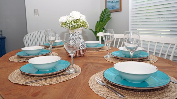 Une table à manger dressée avec des assiettes bleues, des bols blancs et des verres à vin transparents, le tout entouré de sets de table en fibres tressées.