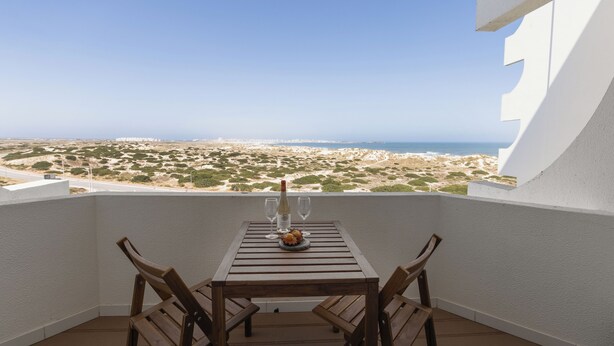 Un balcon avec une table et des chaises en bois, une bouteille de vin et un verre d’eau. La vue comprend une plage et l’océan.