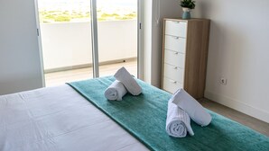 A hotel room with a bed, a green towel on the floor, a wooden cabinet, and a view of the beach through the balcony doors. - Baleal Dream - front of the beach (Peniche)