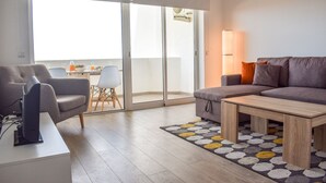 A modern living room with a sofa, a coffee table, and a TV. There is a dining table with chairs and a view through a glass door. - Baleal Dream - front of the beach (Peniche)