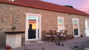 Un edificio de piedra con techo de tejas rojas, una mesa y sillas de madera, y una puerta con un panel de vidrio que refleja un atardecer.