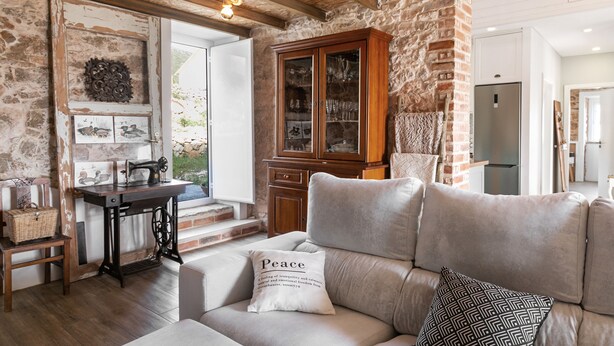 Una sala acogedora con pared de piedra, vigas de madera, un sofá con un cojín que dice 'Peace', una máquina de coser antigua y un aparador con puertas de cristal.