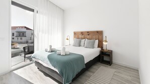 A modern bedroom with a large bed, a wooden headboard, two bedside lamps, and a view through a sliding glass door. - Swell House - Stylish Baleal Beach Apartment (Peniche)