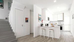 A modern kitchen with white cabinetry, a central island with stools, and a staircase leading to another room. - Swell House - Stylish Baleal Beach Apartment (Peniche)