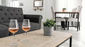 A modern living room with a grey sofa, two wine glasses on a wooden table, a grey potted plant, and a dining area with chairs and a table. - Swell House - Stylish Baleal Beach Apartment (Peniche)