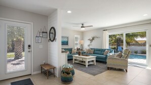 Ein Wohnzimmer mit einem blaugrünen Sofa, einem gemusterten Sessel, einem Couchtisch und einem kleinen Beistelltisch. Eine Tür mit Glasfenster und eine Matte liegen auf dem Boden.